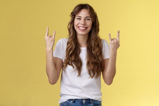 Happy cheerful feminine caucasian girl curly hairstyle show rock-n-roll heavy metal gesture smiling broadly enjoy awesome concert atmosphere stand yellow background joyfully cheering satisfied. Lifestyle.