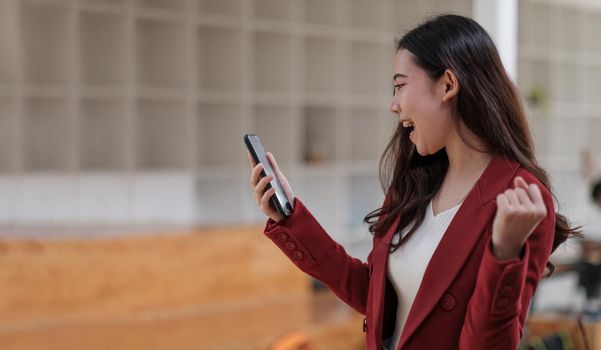 Smiling Asian woman holding mobile phone with fist hand and excited for success in office.