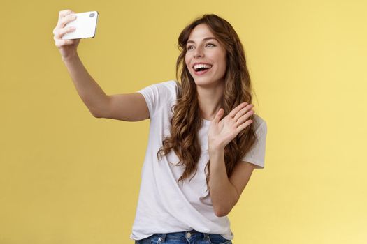 Friendly lively good-looking cheerful feminine girl extend arm hold smartphone record video blog waving front camera smiling broadly talking fans videocalling taking selfie yellow background. Lifestyle.