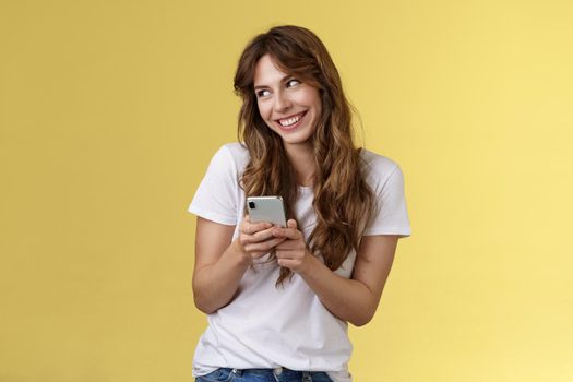 Lovely silly cute flirty girl texting receive romantic lovely gesture look away blushing modest smiling broadly reading bold passionate message stand yellow background joyfully send boyfriend photo. Lifestyle.