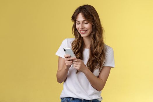 Cheerful lovely girlfriend texting friend pleased cute smile tap smartphone screen smiling broadly look mobile phone display tenderly writing post contemplate touching photo yellow background.