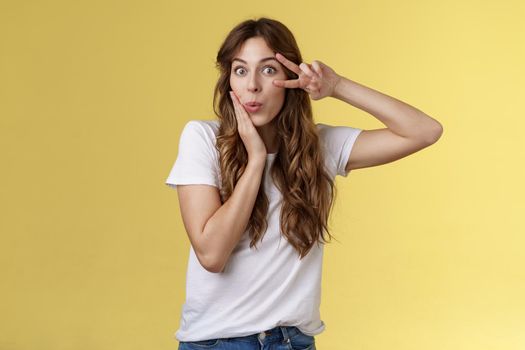 Silly coquesttish good-looking caucasian girl curly hairstyle folding lips amused interested touch cheek feminine sensual pose show victory peace sign upbeat stand intrigued yellow background. Lifestyle.