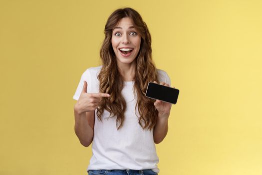 Cheerful surprised cute lucky girl beat best score awesome game showing smartphone display pointing index finger horizontal screen mobile phone introduce cool app smiling broadly yellow background. Lifestyle.