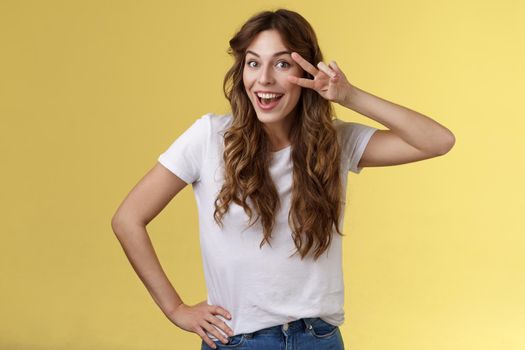 Playful surprised charismatic young caucasian girlfriend having fun look amused enthusiastic show peace victory sign laughing smiling joyfully enjoy summer holidays touch hip stand yellow background. Lifestyle.