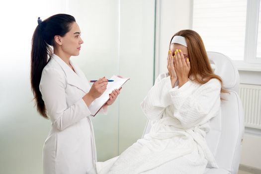 Cosmetologist consulting a female patient and making notes during medical appointment in modern beauty clinic