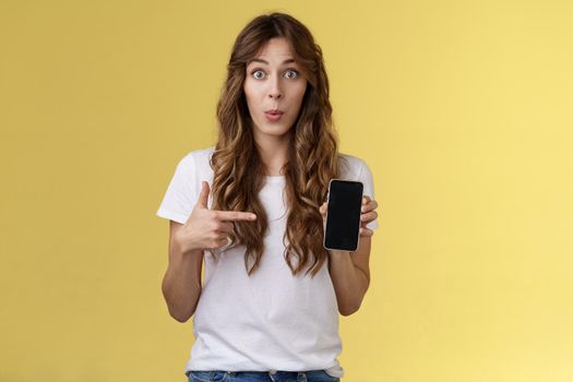 Intriguing app check out. Enthusiastic surprised attractive girlfriend gossiping friend new boyfriend showing curious photo smartphone hold mobile phone pointing telephone screen yellow background.