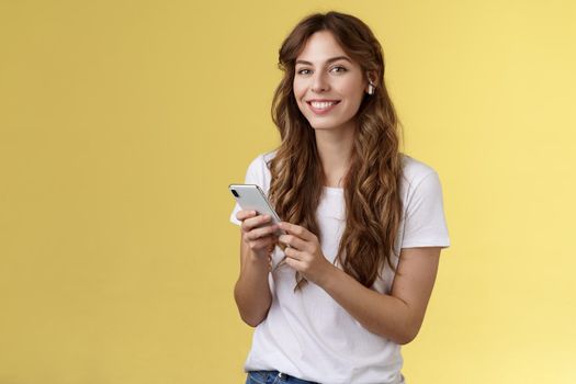 Feminine lovely curly-haired woman white t-shirt hold smartphone picked awesome new song listen music wearing wireless earphones smiling delighted camera enjoy earbuds beats yellow background. Lifestyle.