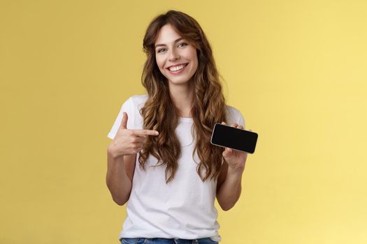 Cheerful good-looking friendly carefree young girl hold smartphone horizontal pointing index finger mobile phone screen smiling broadly introduce awesome game app stand yellow background.