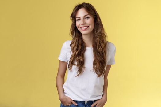 Attractive carefree confident friendly-looking charming feminine ubran woman long curly hair smiling toothy pleased tilt head intrigued hold hands pockets self-assured pose stand yellow background. Lifestyle.