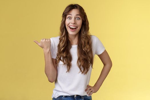 So amazing lets go together. Lively excited european female curly haircut wanna visit cool party smiling joyfully pointing thumb left grinning thrilled look camera admiration yellow background.