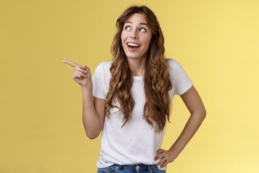 Wondered impressed charismatic fascinated smiling happy girl pointing look upper left corner speechless surprised grinning toothy happiness joy expression contemplate great view yellow background.