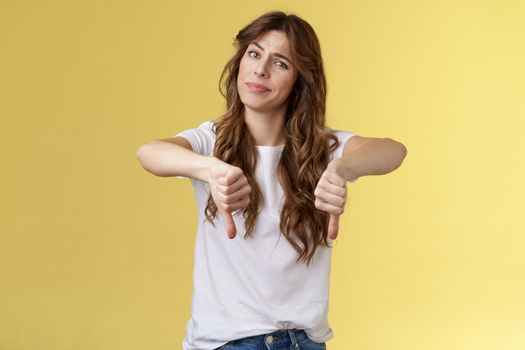 Honestly very bad. Disappointed upset curly-haired girl tilt head smirk displeased disagree show thumbs down sign dislike unimpressed awful idea stand yellow background uninterested.