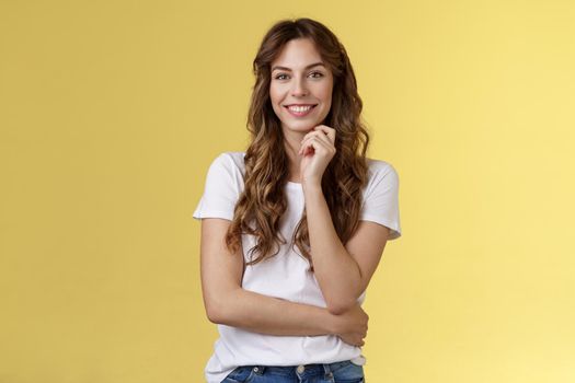 Intrigued attractive european brunette curly long hair look interested listen curious suggestion touch cheek interest temptation smiling toothy pleased good conversation stand yellow background.