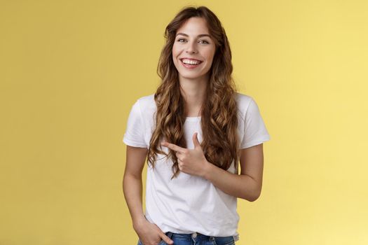 Outgoing charming happy pretty woman curly long hairstyle pointing forefinger left indicate advertisement laughing carefree satisfied hold hand pocket casual friendly pose yellow background.