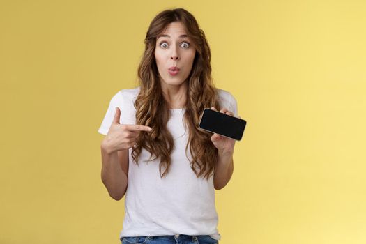 Surprised enthsuastic intrigued cute curly-haired caucasian girl folding lips amused curiously pointing smartphone screen stare camera ambushed astonished describe cool mobile phone app.
