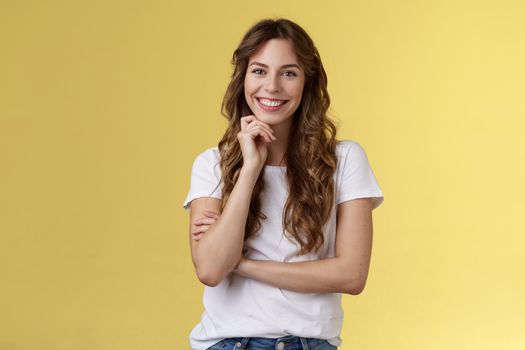 Tender feminine enthusiastic charming european woman curly long hairstyle laughing silly gaze grinning joyfully touch chin intrigued standing curious interested listening amused yellow background.