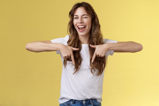 Hey visit my link would not regret. Lively cheeky enthusiastic woman curly long haircut wink sassy smiling broadly show you perfect copy space pointing down bottom promo yellow background.