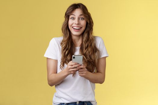 Extremely happy enthusiastic surprised joyful lovely feminine girl receive pleasant touching message online hold smartphone smiling broadly look camera joyful amused yellow background.