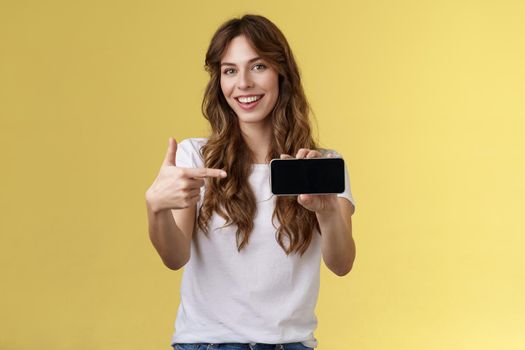 Confident sassy good-looking stylish woman introduce awesome game app show smartphone pointing index finger blank mobile phone screen smiling delighted recommend try application yellow background. Lifestyle.