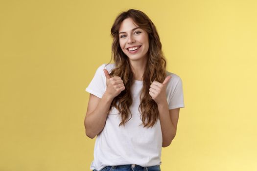 Good have my support. Cheerful positive cute lively curly-haired european girl show thumbs-up approval like gesture tilt head pleased accepting judging fine choice stand yellow background.