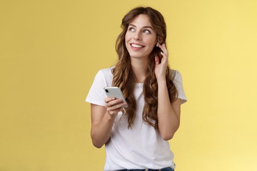 Dreamy happy cheerful curly-haired girl look around contemplate beautiful summer weather listening music touch wireless earbud calling friend talking via earphones hold smartphone yellow background. Lifestyle.