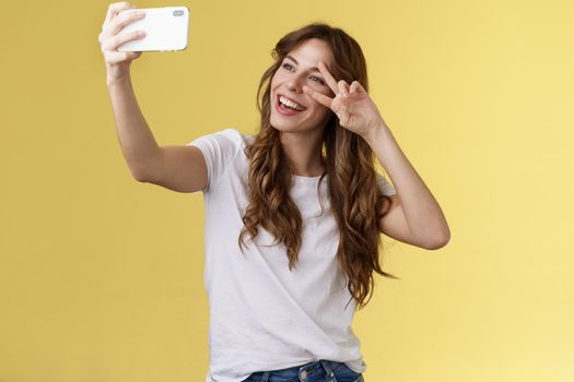 Joyful charismatic good-looking curly-haired woman white t-shirt show peace victory sign smiling broadly posing tender friendly expression smartphone front camera take selfie yellow background. Lifestyle.