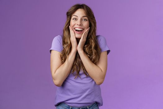 Happy lively lucky enthusiastic girl curly hairstyle open mouth admiration fascinated smiling broadly touch cheek impressed surprised awesome incredible positive news stand purple background.