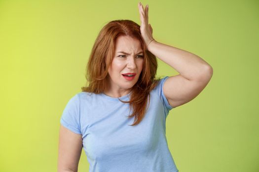 Something important slip my mind. Concerned worried upset redhead mature woman punch forehead turn away frustrated frowning disappointed forget cancel appointment stand green background.