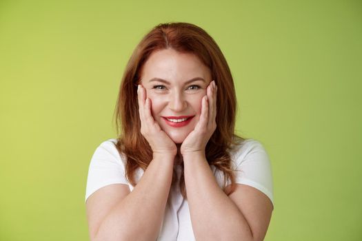 Happy delighted cheerful lucky redhead middle-aged caucasian woman. blushing joyfully receive touching cute gift touch cheeks pleased delighted smiling broadly feel happiness joy green background.