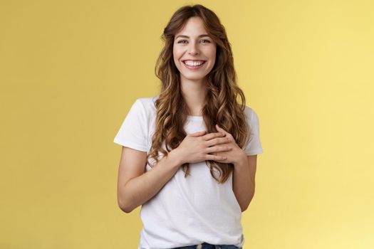 Tender heartwarming kind gentle feminine girl curly long hairstyle look sincere delighted squinting joyfully smiling broadly touch heart hold hands chest grateful lovely gaze stand yellow background. Lifestyle.