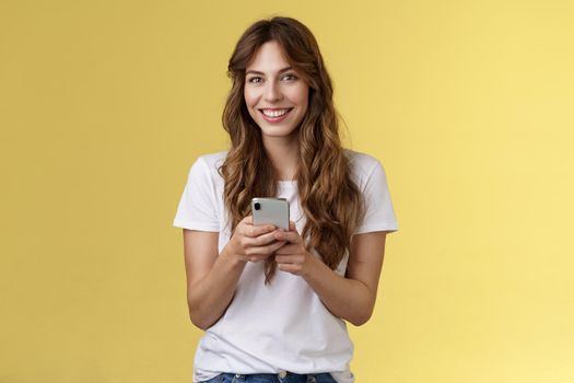 Enthusiastic charming sociable young girl messaging friend sending photos social media hold smartphone look camera happily friendly smiling stand yellow background casual outfit. Technology concept