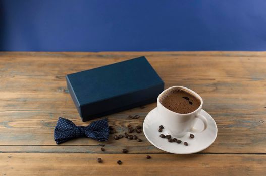 White ceramic cup of coffee with bow tie and gift box on old wooden table with some beans. Father day card.
