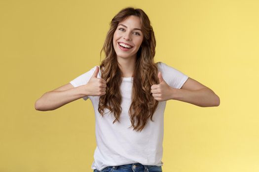 Outgoing positive confident lively young girl long curly haircut support great effor show thumbs up positive reply approving good choice like your outfit smiling broadly yellow background. Lifestyle.