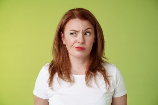 Perplexed doubtful unsure redhead wife smirking unsatisfied frowning look aside displeased disappointed hesitating pondering strange advice thoughtfully stare away stand green background.