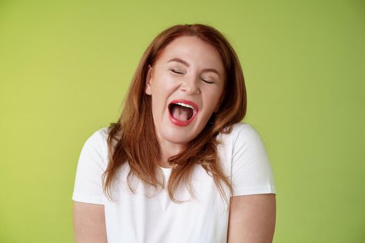 Lazy weekends finally time sleep. Cheerful redhead middle-aged 50s woman yawning satisfied close eyes feel sleepy wake up early morning wanna take nap stand green background watching boring film.