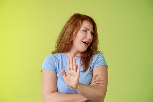 Enough wasting my time. Ignorant uninterested redhead mature woman turn away displeased reluctant show stop no gesture hold palm refusal rejecting unpleasant offer green background.