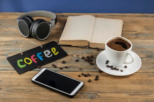A cup of coffee, headphone, open book and a board with the inscription "coffee" written in a colorful alphabet. Coffee break, the concept of waking up in the morning.