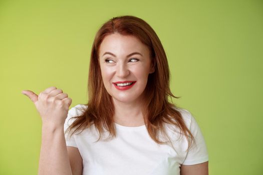 Intrigued charismatic redhead middle-aged woman smiling temtation interest pointing looking left curiously check-out cool promo share awesome place location stand green background.