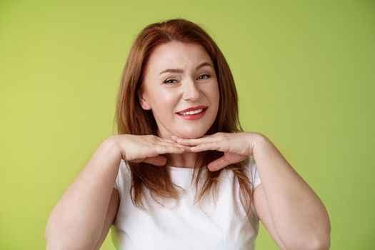 Looking good. Happy cheerful redhead middle-aged 50s woman smiling delighted hold hands under chin accept flaws blemished like own skin condition apply aging creme cosmetics green background.