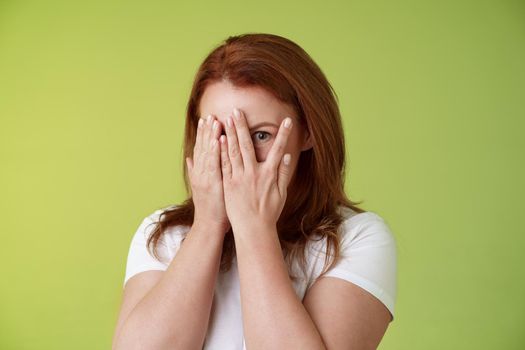 Not peeking. Playful charismatic middle-aged ginger redhead woman close face palms look through fingers one eye stare camera intrigued waiting surprise gift stand green background.