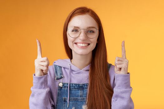 Lively charismatic pleasant redhead female student helpful pointing up index fingers showing awesome promo offer smiling delighted white teeth indicating advertisement recommend use product.