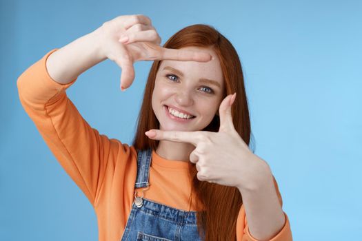 Joyful attractive sincere redhead young girl searching inspiration find perfect angle take good shot make hand frames look through delighted amused smiling broadly white teeth, blue background.