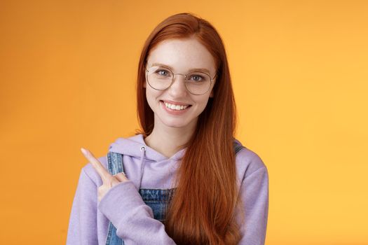 Friendly good-looking modern redhead young girl pointing left index finger showing awesome place suggesting go hang out smiling joyfully casually talking discuss new product, orange background.