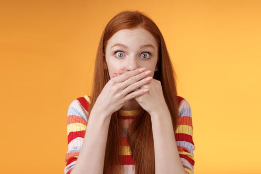 Oops sorry. Girl feeling awkward saying inappropriate word standing unconfident cover mouth palms wide eyes staring camera shocked express surprise nervously standing orange background.