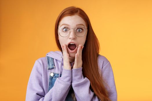 Shocked impressed redhead girlfriend drop jaw gasping screaming wow omg touch cheek near mouth wide eyes surprised reacting astonishing news fresh rumors gossiping amazed, orange background.