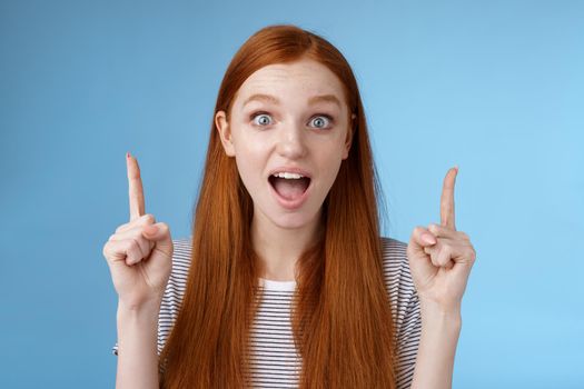 Surprised happy enthusiastic young redhead woman reacting impressed upper hanging promo pointing up index fingers drop jaw amused look thrilled camera telling about awesome offer, blue background.