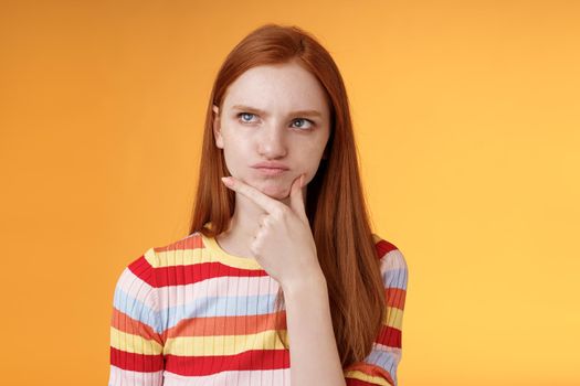 Perplexed hesitant serious-looking ginger girl blue eyes weihing choices pouting touch chin look upper left corner thoughtful thinking puzzled solution, pondering decision, orange background.