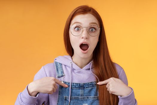 Lifestyle. Shocked emotive surprised redhead european woman wearing glasses hoodie drop jaw gasping stunned pointing herself full disbelief cannot believe win being picked look impressed orange background.