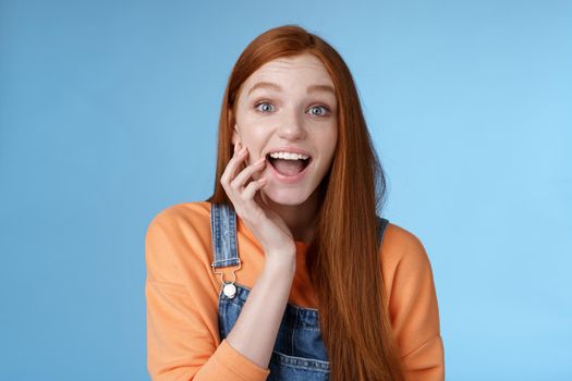 Excited enthusiasitc charismatic redhead female friend look surprised amused touch cheek open mouth thrilled discussing future prom rejoicing standing blue background joyful delighted.