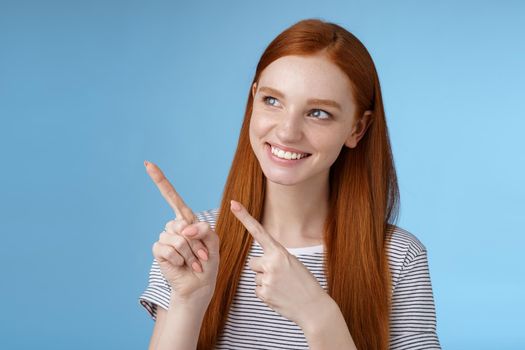 Attractive redhead female model pure clean skin ginger long hair pointing looking left intrigued smiling satisfied indicating interesting advertisement intrigued using new product, blue background.